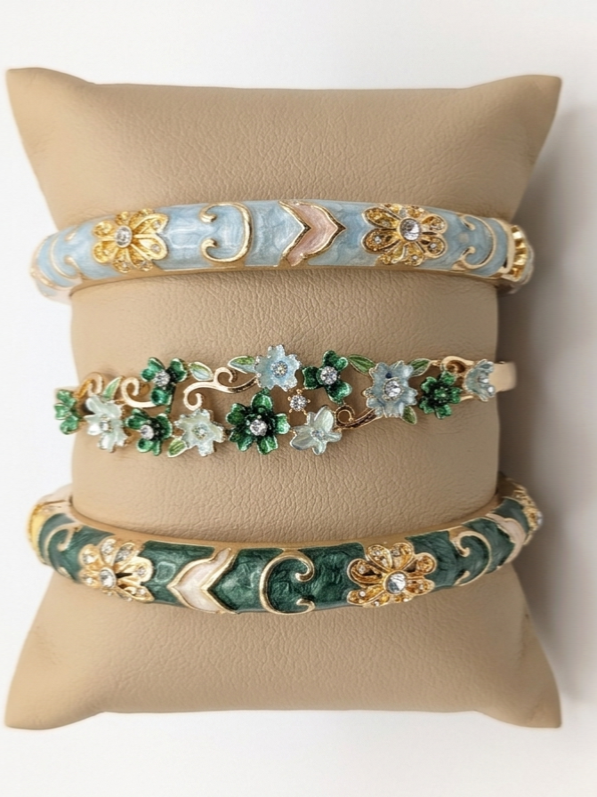 Composition de trois bracelets élégants sur un coussin en lin, mettant en valeur un bracelet central orné de délicates fleurs émaillées vertes et bleu pastel avec des touches de strass.