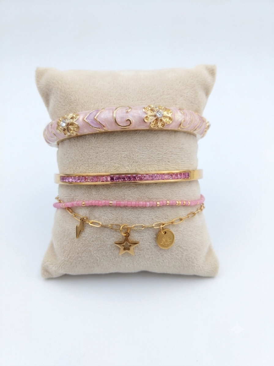Bracelet jonc romantique pour femme en émail rose poudré avec détails floraux dorés, présenté sur un coussin en lin avec une composition de bracelets roses et dorés.