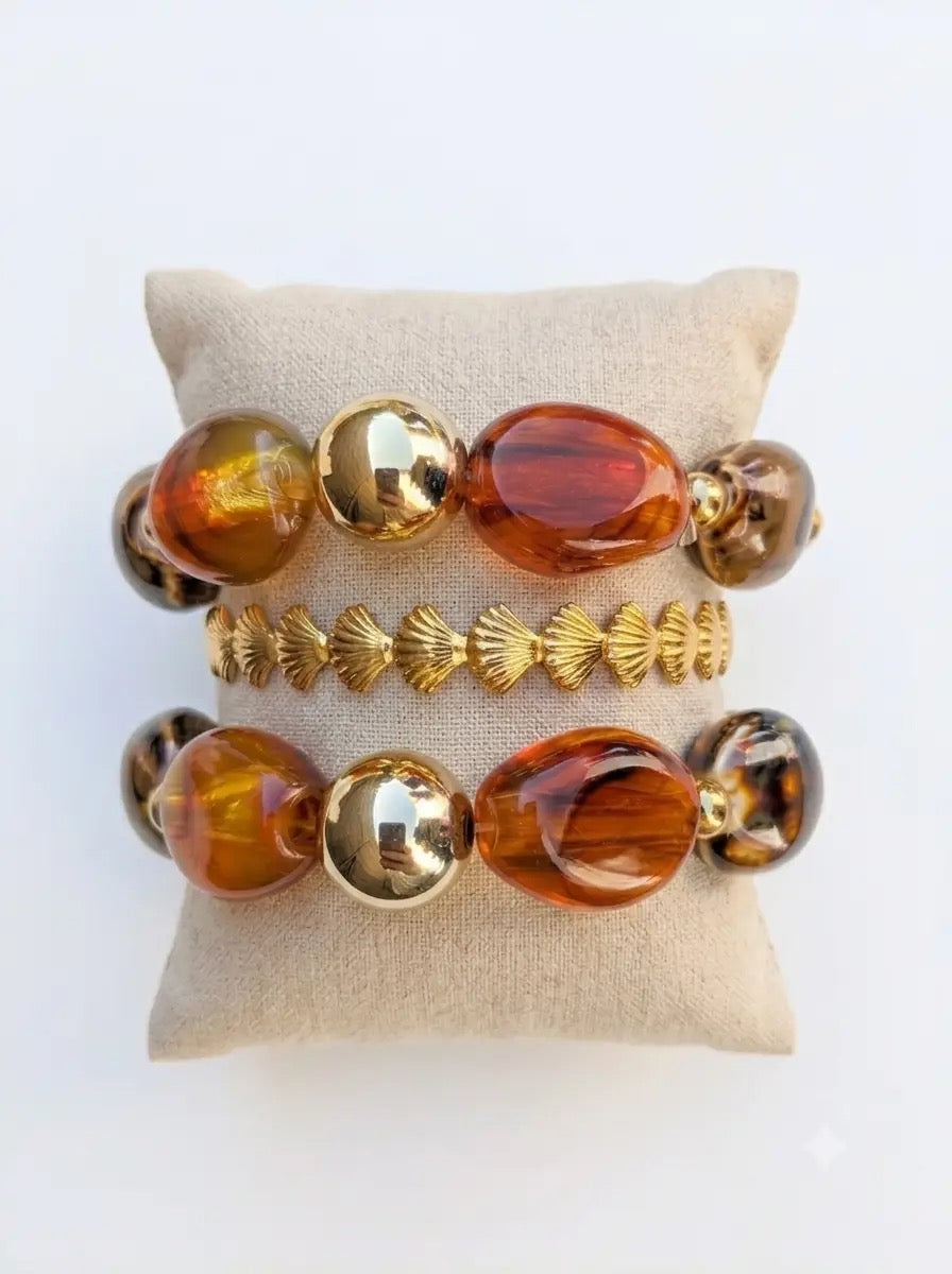 Bracelets élégants avec grosses perles effet ambre écaille de tortue et sphères dorées, en acier inoxydable, présentés sur un coussin en lin.